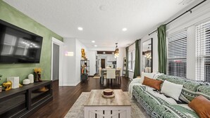 A modern living room with a sofa, a coffee table, and a TV. There is a dining area with a table and chairs. - Space Cowboy Retreat • Sleeps 11 • Fire Pit • Near NRG (Houston)