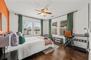 A room with a bed, a desk, a chair, a ceiling fan, and large windows with curtains.