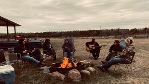 Eine Gruppe von Menschen sitzt an einem Lagerfeuer in einer natürlichen Umgebung, mit einem grasbewachsen Feld und Bäumen im Hintergrund.