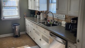 A kitchen with white cabinets, a stainless steel dishwasher, a ceiling fan, and a window with blinds.