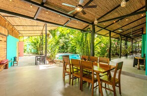 A covered outdoor dining area with a pool, wooden furniture, and tropical plants.