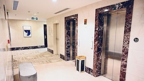 A modern elevator area with marble walls, a bench, and a small trash bin. - Golden Hospitality Hotel (Makkah)