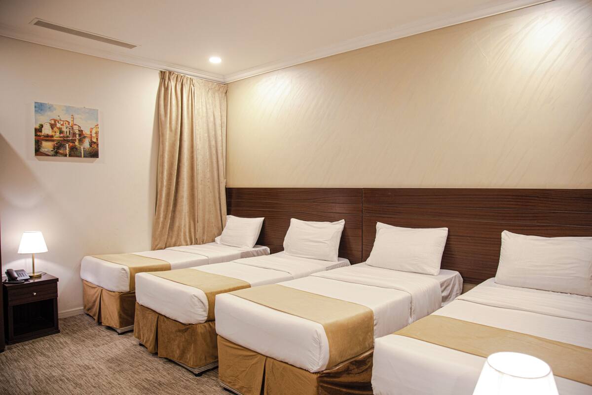A hotel room with four single beds, a wooden headboard, a painting on the wall, and a nightstand with a lamp.