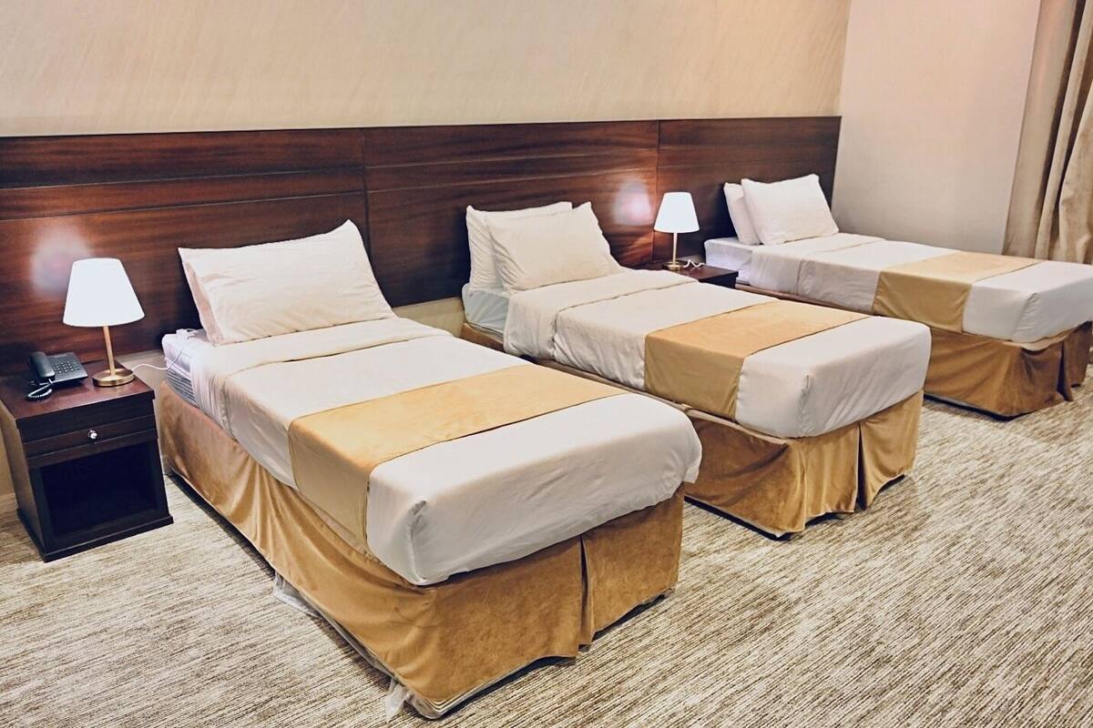 A hotel room with three single beds, each with a bedside table and lamp.