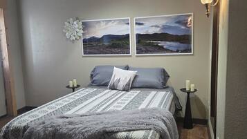 Una cama bien hecha con ropa de cama estampada, dos cuadros de paisajes colgados en la pared, una mesita con velas e iluminación colgante.