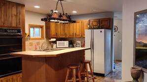 Une cuisine avec des meubles en bois, un îlot central doté de tabourets, un réfrigérateur, un four à micro-ondes et un grand tableau encadré au mur.