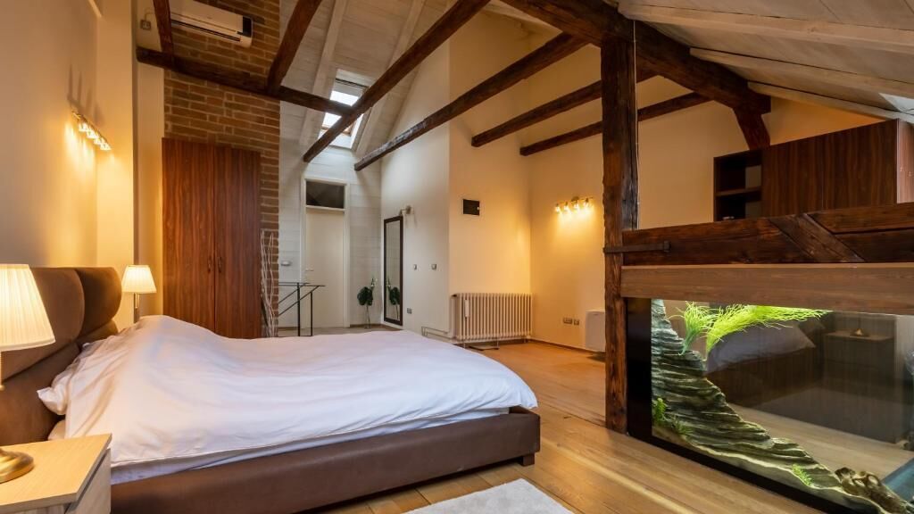 A bedroom with a large bed, wooden beams, a glass fish tank, and a view of a wooden structure.