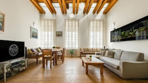 A spacious living room with a wooden ceiling, large windows, a flat-screen TV, a sofa, a coffee table, and a dining area with chairs and a table.
