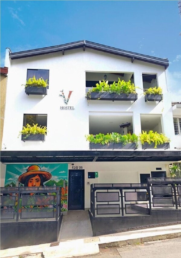 A white building with the word "HOSTEL" on the facade, balconies with plants, and a mural of a woman with a hat. - Vida Hostel (Medellín)