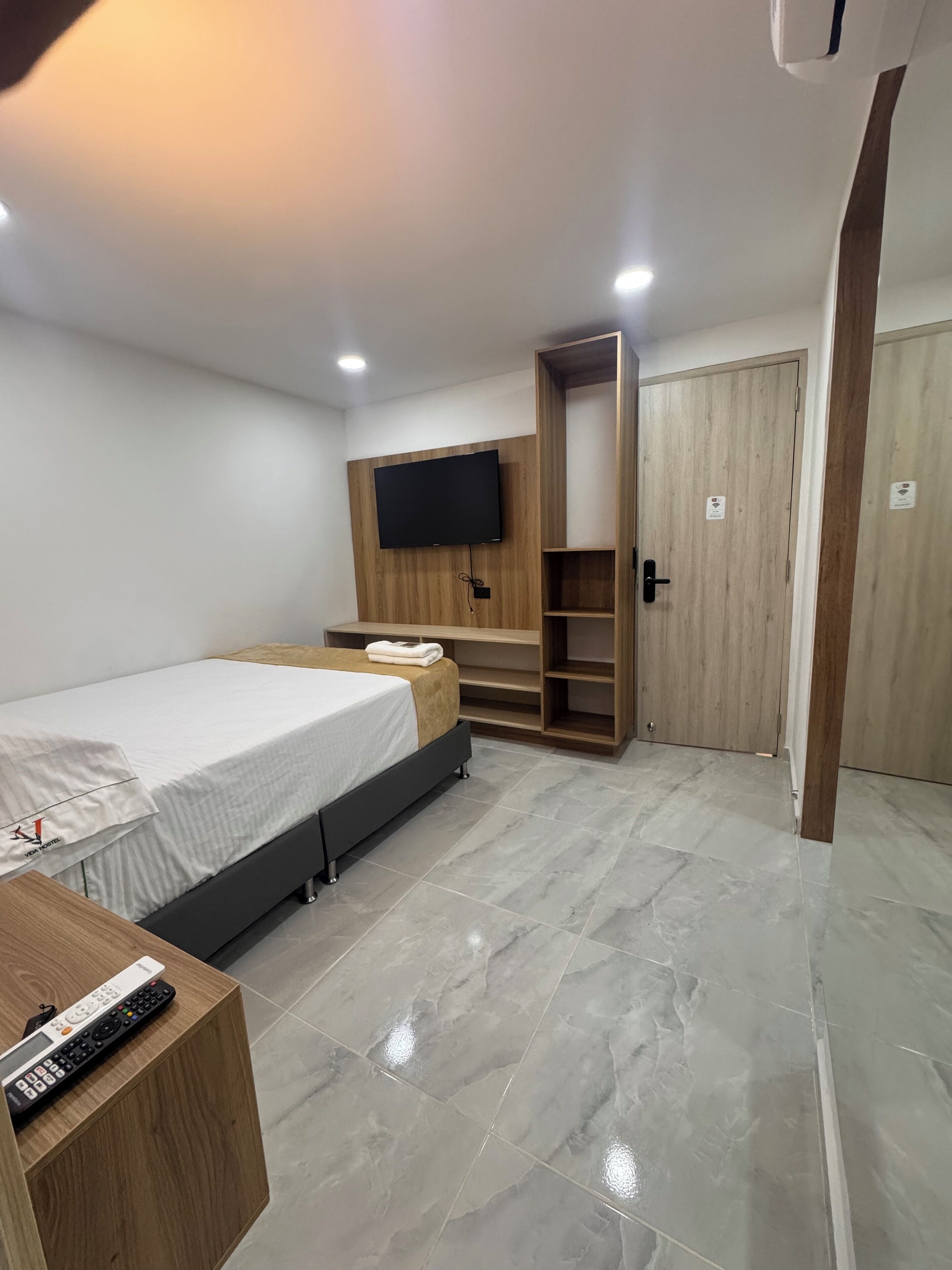 Une chambre moderne avec un lit, un téléviseur fixé au mur en bois, une armoire en bois et une petite table avec une télécommande.