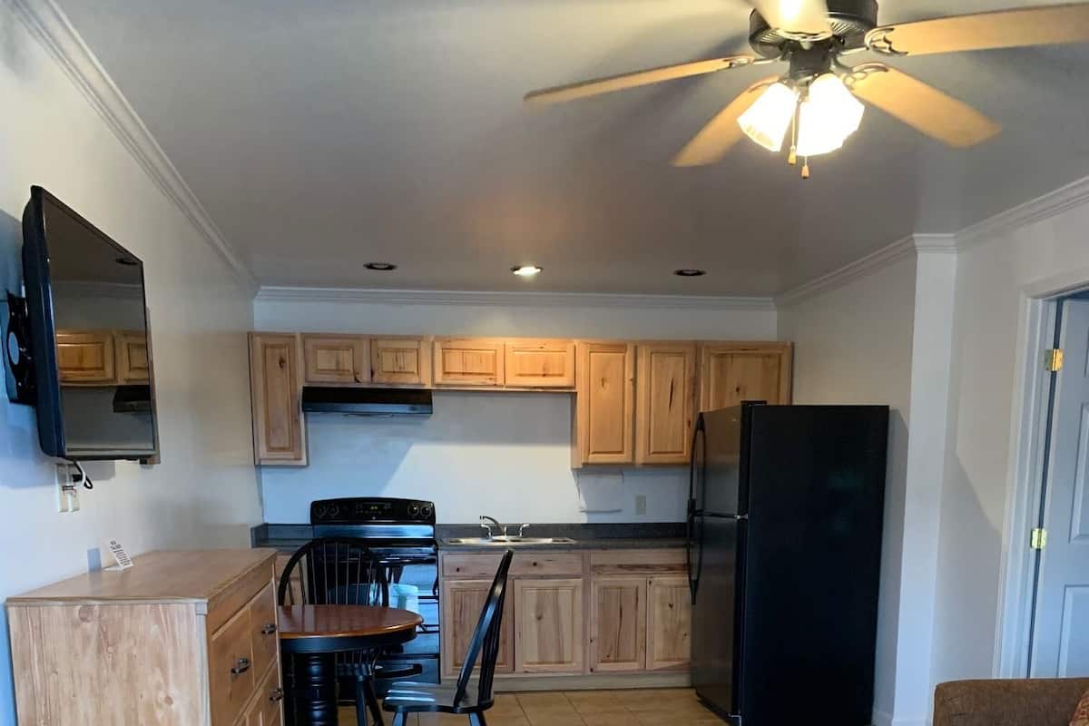 A compact kitchen with wooden cabinets, a black refrigerator, a dining table with chairs, and a ceiling fan.