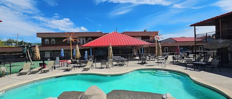 Ein Swimmingpool mit Liegestühlen und Sonnenschirmen, umgeben von einem eingezäunten Bereich, im Hintergrund ein Gebäude.
