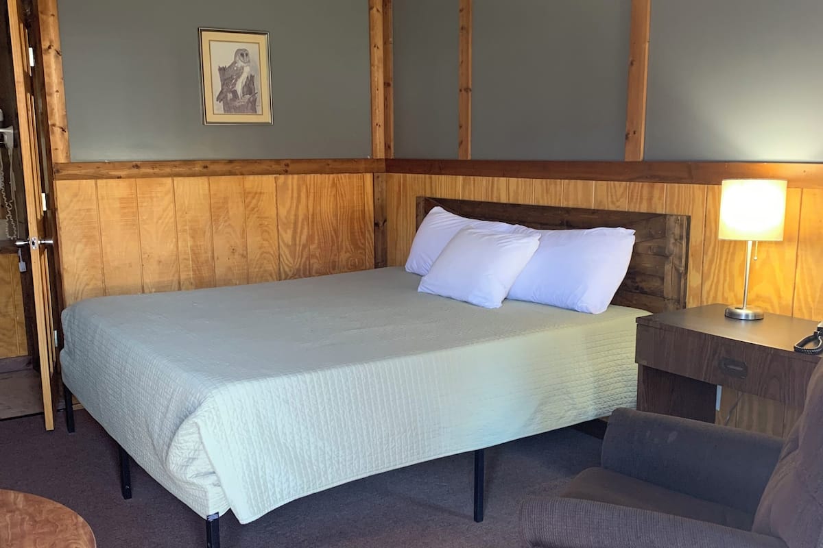 A bedroom with a bed, a nightstand, a lamp, a chair, and a framed picture on the wall.