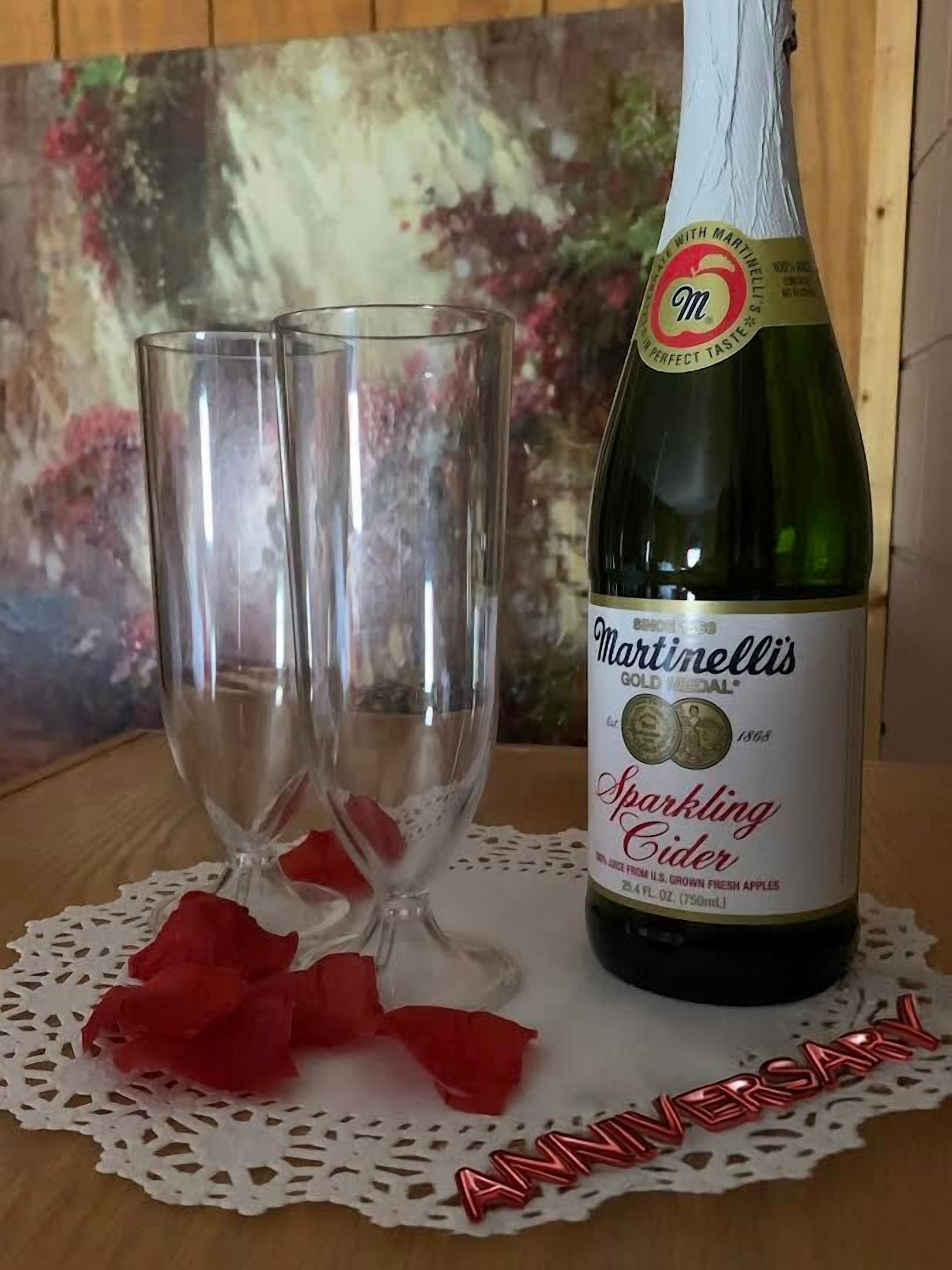 A bottle of Martinellis Sparkling Cider and two empty glasses on a doily with red rose petals.