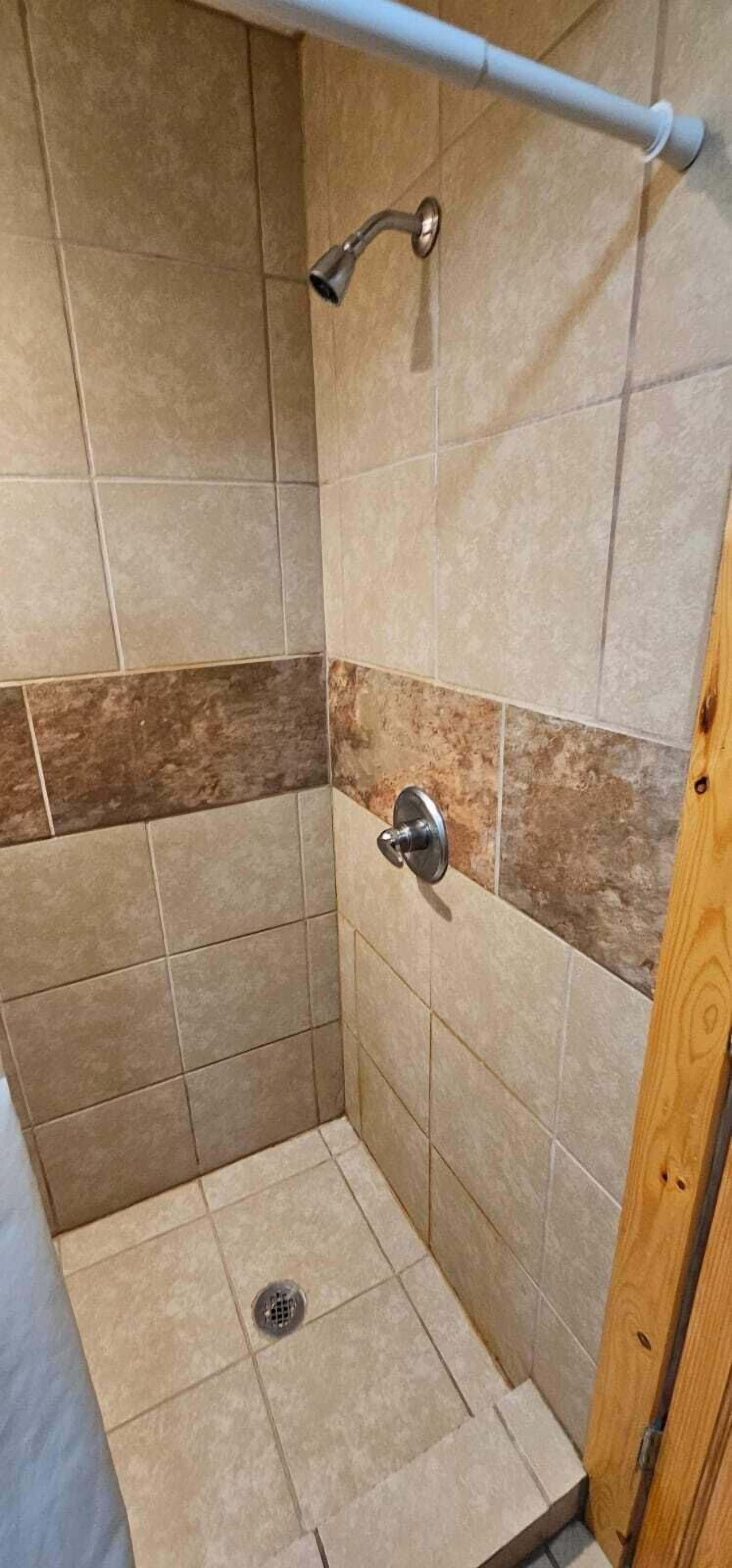 A shower stall with beige tiles, a showerhead, and a wooden door frame.