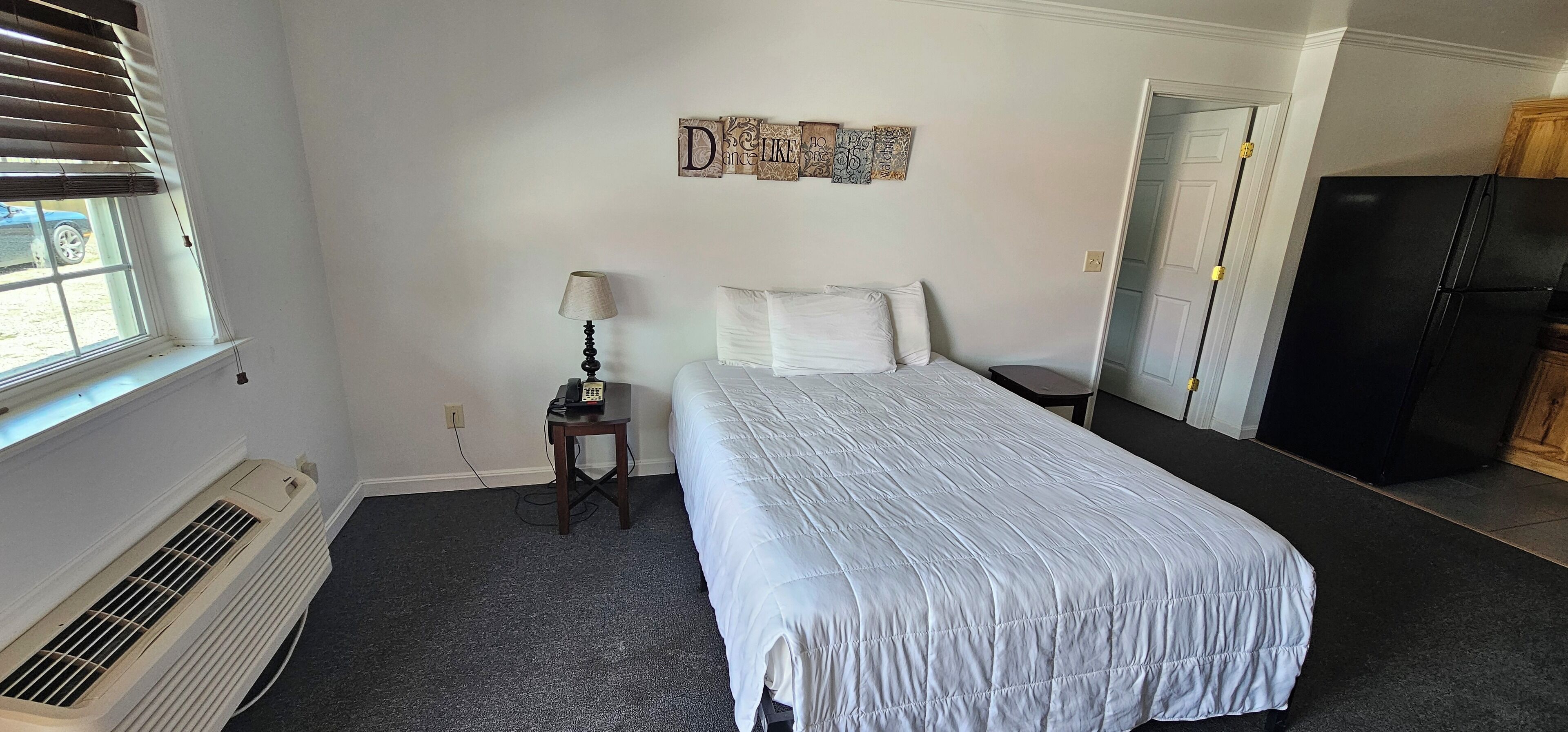 A bedroom with a bed, a bedside table with a lamp, a wall decoration, a window, and a door leading to a kitchen.