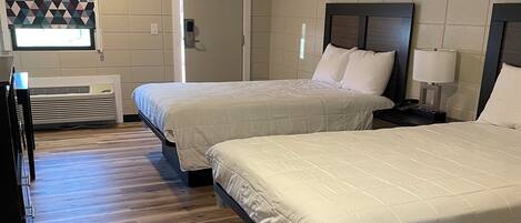 A hotel room with two beds, a wooden floor, a wall-mounted air conditioner, and a window with a geometric pattern.