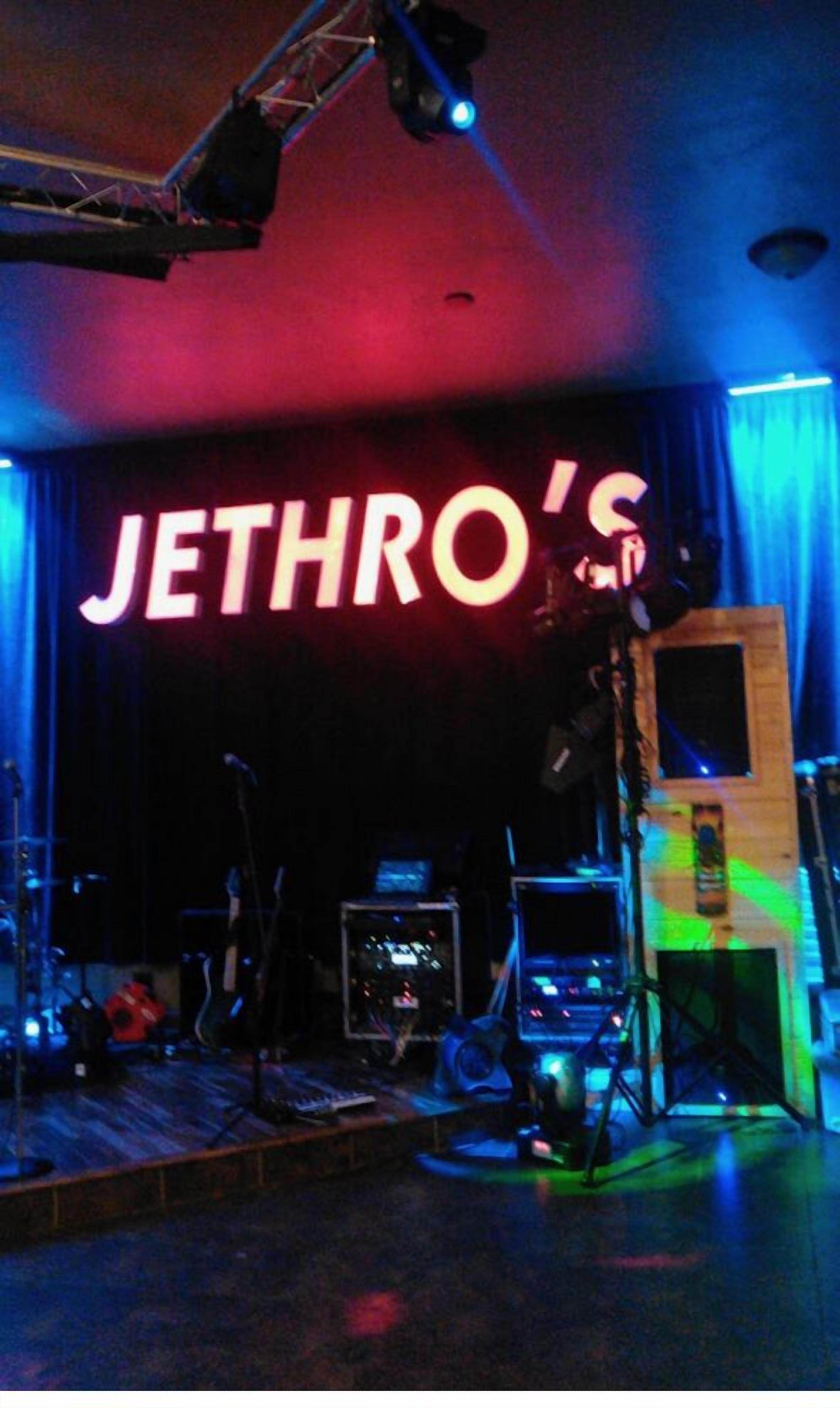 Un escenario con un gran letrero iluminado que dice "JETHRO'S" y diverso equipo musical.