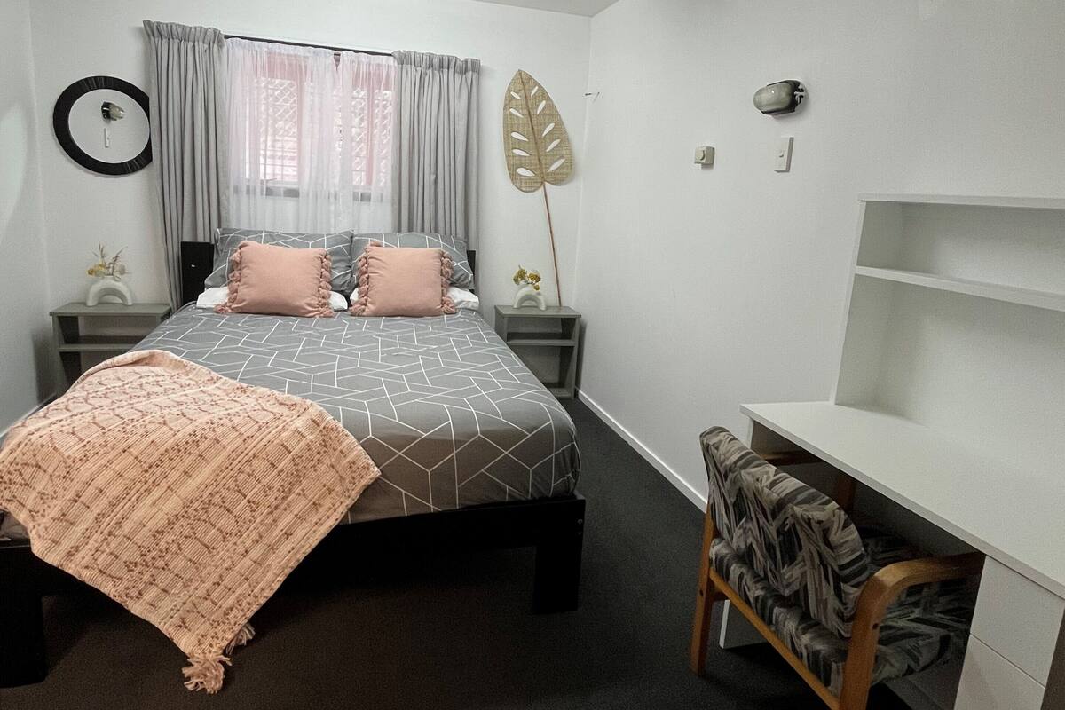 Une chambre à coucher comprenant un lit, des tables de chevet, un bureau avec une chaise, une grande feuille décorative, un miroir et une fenêtre avec des rideaux.