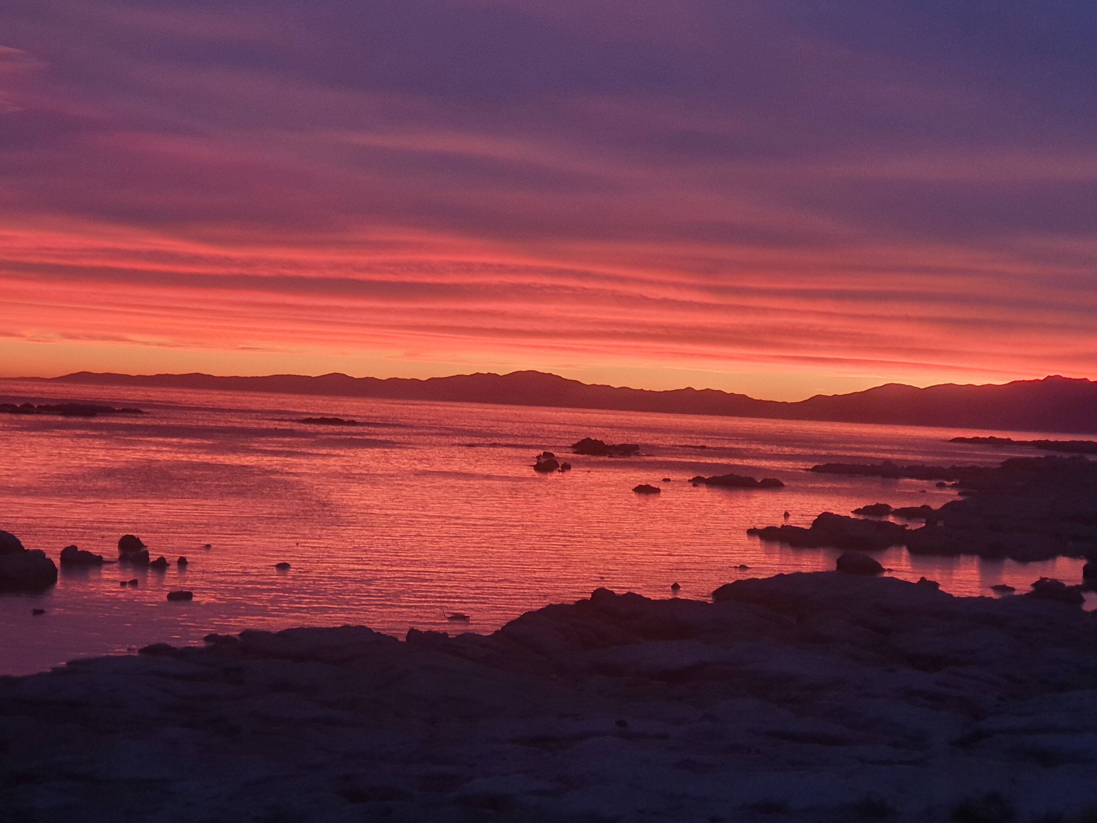 Un paisaje costero con un atardecer sobre el agua, costa rocosa y montañas a lo lejos.
