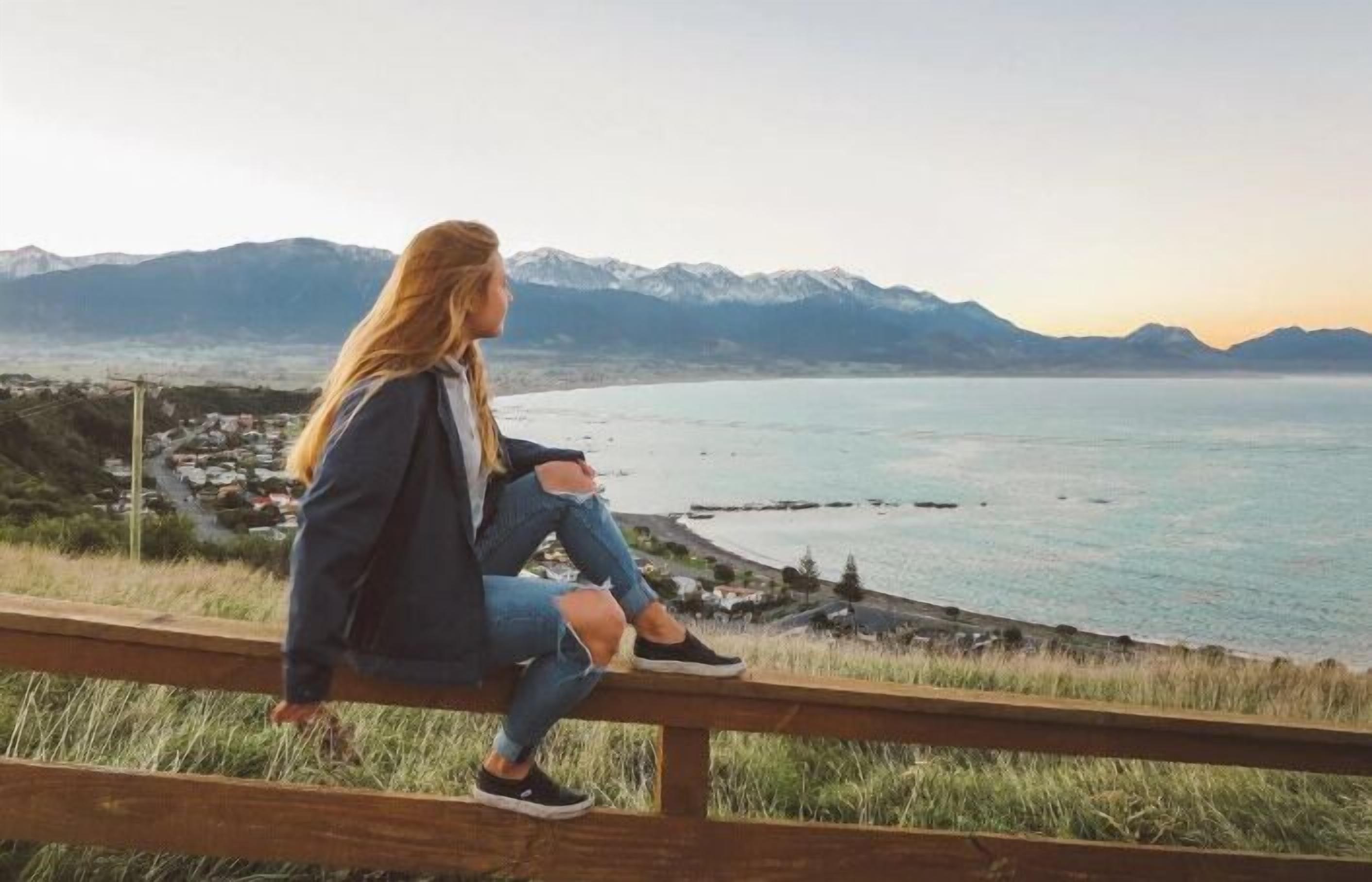 A person sitting on a wooden bench overlooking a coastal landscape with mountains in the background.