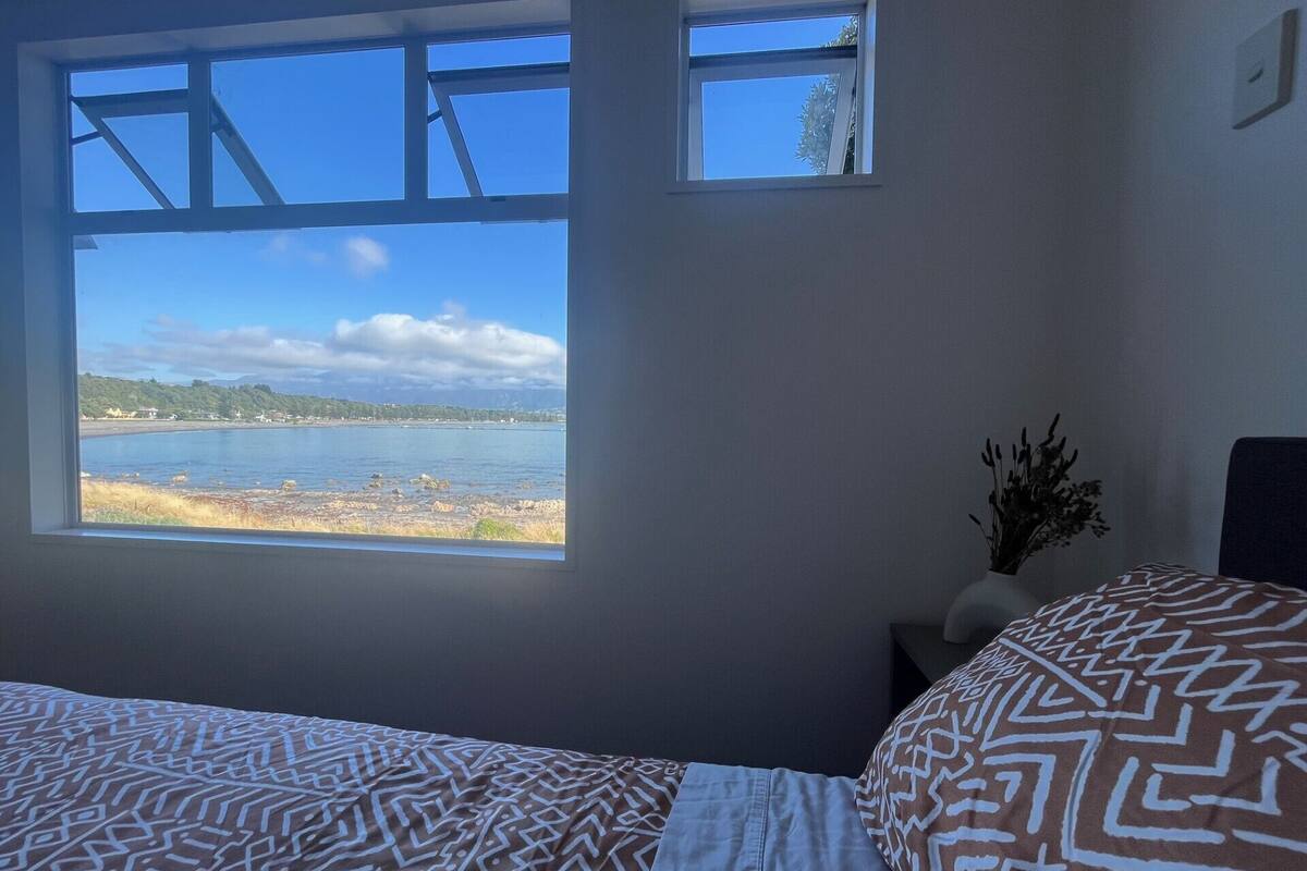 Une chambre avec une grande fenêtre donnant sur une plage et des montagnes. On y trouve un lit avec une literie à motifs et une petite plante posée sur une table de chevet.