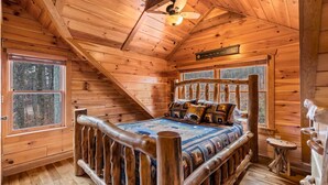 Een slaapkamer in een blokhut met een houten bed, een plafondventilator en een raam met uitzicht op bomen.
