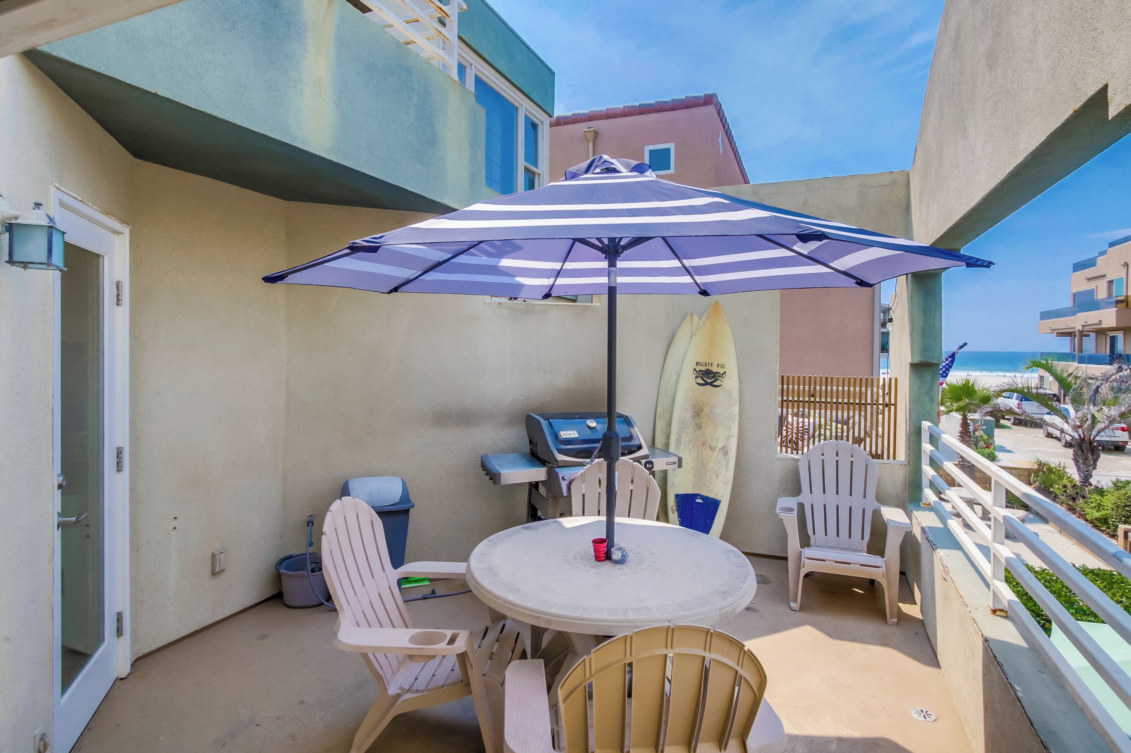 A balcony with a round table, chairs, a striped umbrella, a surfboard, and a grill.