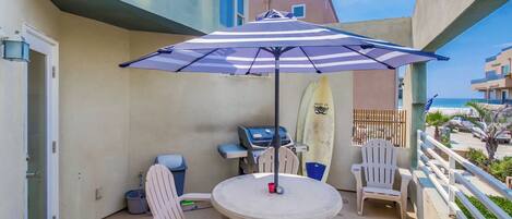 A balcony with a round table, chairs, a striped umbrella, a surfboard, and a grill.