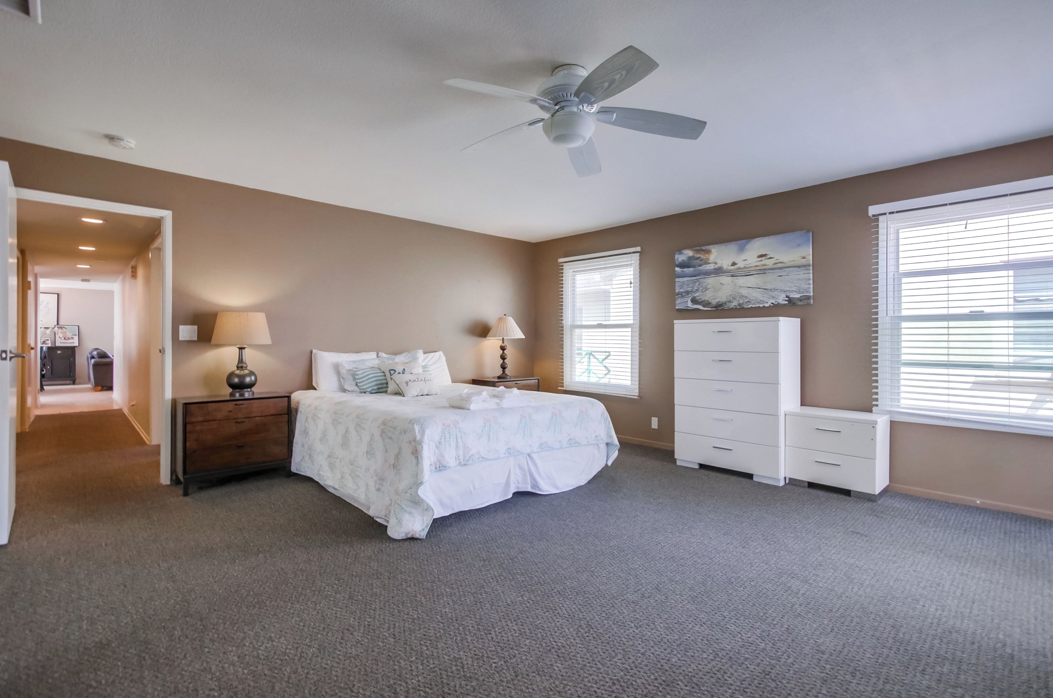 Une chambre à coucher comprenant un lit, des tables de chevet, une commode, un ventilateur de plafond et un tableau accroché au mur.