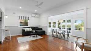 A spacious living area with a black sofa, a dining table with chairs, and large windows offering a view of greenery. - Charming Cottage by the Water Pet Friendly WoyWoy (Woy Woy)