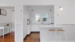 A modern kitchen with white cabinetry, a wooden floor, and a bar area with stools. - Charming Cottage by the Water Pet Friendly WoyWoy (Woy Woy)