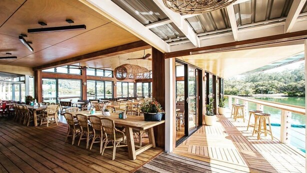 A spacious indoor dining area with wooden flooring and tables, large windows, and a view of a river. - Charming Cottage by the Water Pet Friendly WoyWoy (Woy Woy)