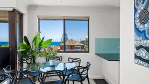 A dining area with a glass table and chairs, a potted plant, and a view of the sea and buildings through large windows. - Dream View Steps from the Beach (Coogee)