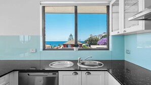 A kitchen with a window view of a coastal town, a stainless steel sink, and a dishwasher. - Dream View Steps from the Beach (Coogee)