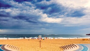 A beach with a curved pathway leading to the water, a lifeguard sign, and people walking along the shore. - Dream View Steps from the Beach (Coogee)