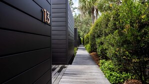 Un étroit sentier bordé de murs recouverts d’un bardage sombre et d’une végétation luxuriante, qui mène à l’entrée d’un bâtiment.