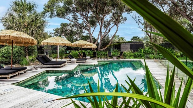Un espace piscine doté de chaises longues et de parasols en chaume, entouré d’une végétation luxuriante.
