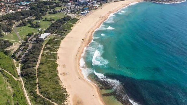 An aerial view of a coastal area with a sandy beach, clear blue ocean, and surrounding greenery.