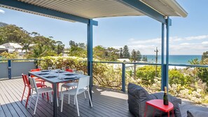 A covered outdoor deck with a table set for dining, overlooking a scenic coastal view. - Oceanview Escape One Hour from Sydney (Wombarra)