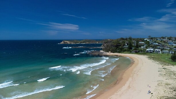 Une plage côtière aux eaux limpides, avec un rivage sablonneux et une zone résidentielle bordée de maisons.