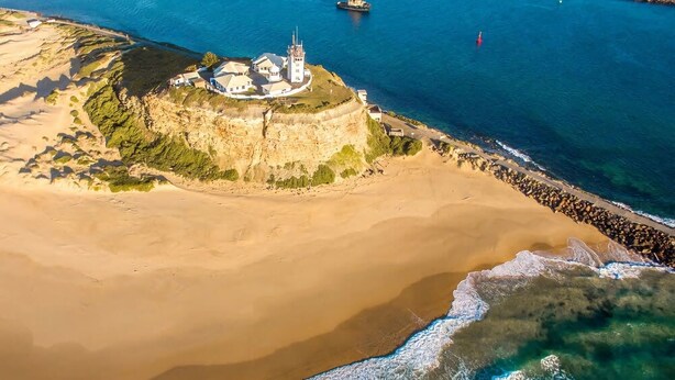 Vue aérienne d’une zone côtière, avec un imposant bâtiment blanc doté d’une tour, entouré d’une plage de sable et de falaises rocheuses.