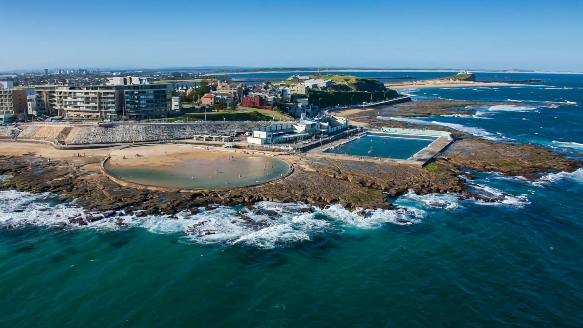 An aerial view of a coastal area with a beach, rocky shoreline, and a large building complex.