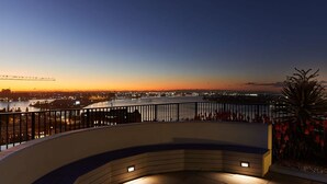 Une terrasse au bord de l’eau, dotée d’une balustrade incurvée, qui offre une vue sur le paysage urbain au crépuscule.