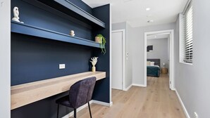 Un intérieur moderne avec un mur d’accent bleu foncé, un bureau en bois, une chaise foncée et une vue sur une chambre à coucher avec un lit et des stores aux fenêtres.