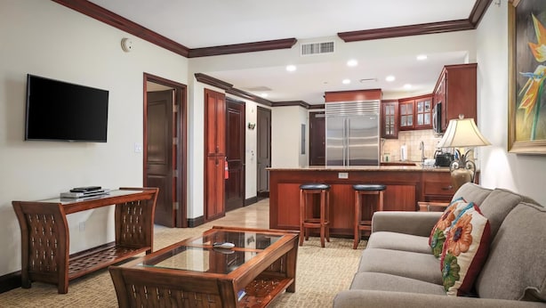 A living room with a sofa, a coffee table, a TV, and a kitchen area with a fridge and bar stools.