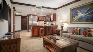 A modern kitchen with wooden cabinets, a central island with stools, a dining area with a glass-top table, and a living room with a sofa and a painting.