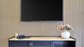 Un salón moderno con televisor de pantalla plana montado en una pared de madera, una consola de madera con adornos y un jarrón con flores.