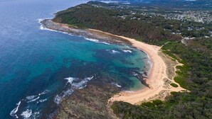 An aerial view of a coastal area with a beach, forested hills, and a distant town. - The Forrester Beachside Retreat Resort Style Pool Oasis (Forresters Beach)