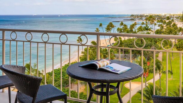 A balcony with a view of the ocean and beach, featuring wicker furniture and a magazine. - Waikiki Shore 906 Ocean View 1 Bedroom (Honolulu)
